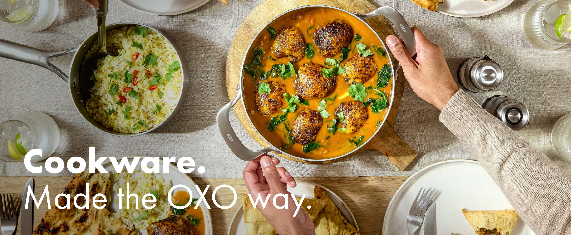 Overhead view of an OXO stainless steel sauté pan with meatballs in a rich sauce at a shared table with rice, naan and drinks, with hands serving.