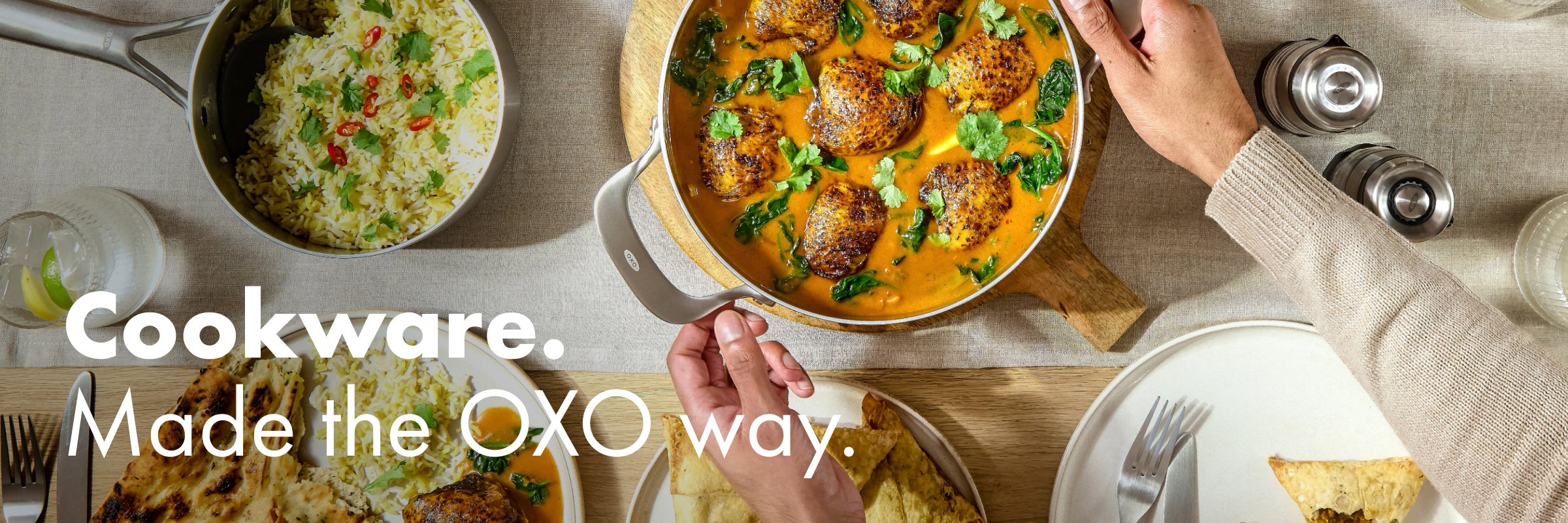 Overhead view of an OXO stainless steel sauté pan with meatballs in a rich sauce at a shared table with rice, naan and drinks, with hands serving.