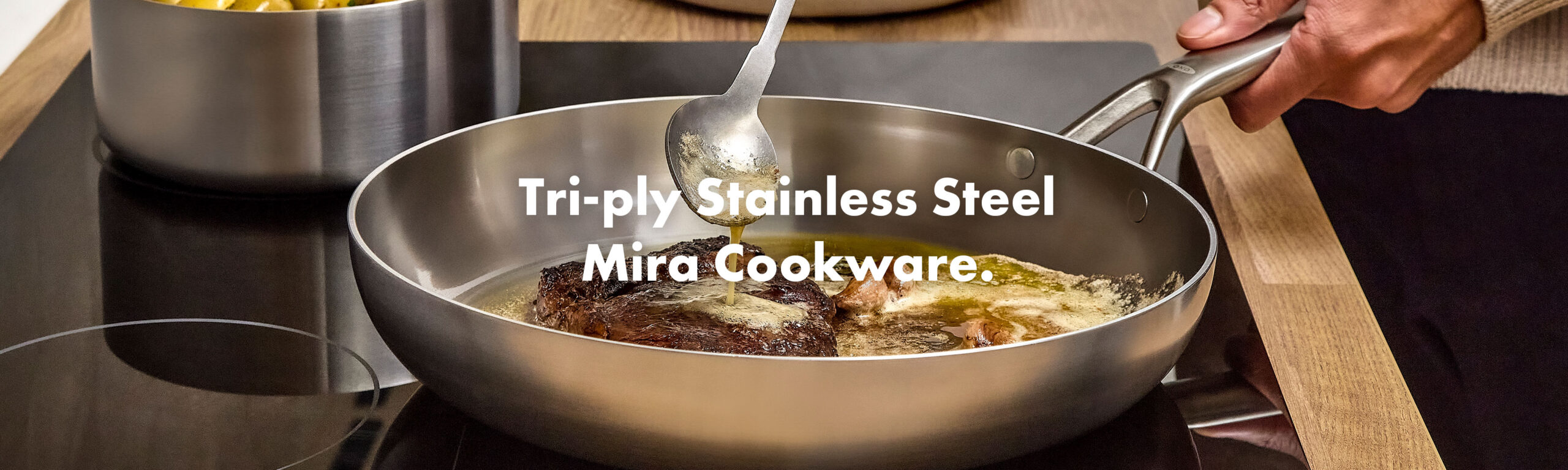 Basting a seared steak with butter in an OXO Mira tri‑ply stainless steel frying pan on an induction hob, with a matching saucepan of potatoes in the background.